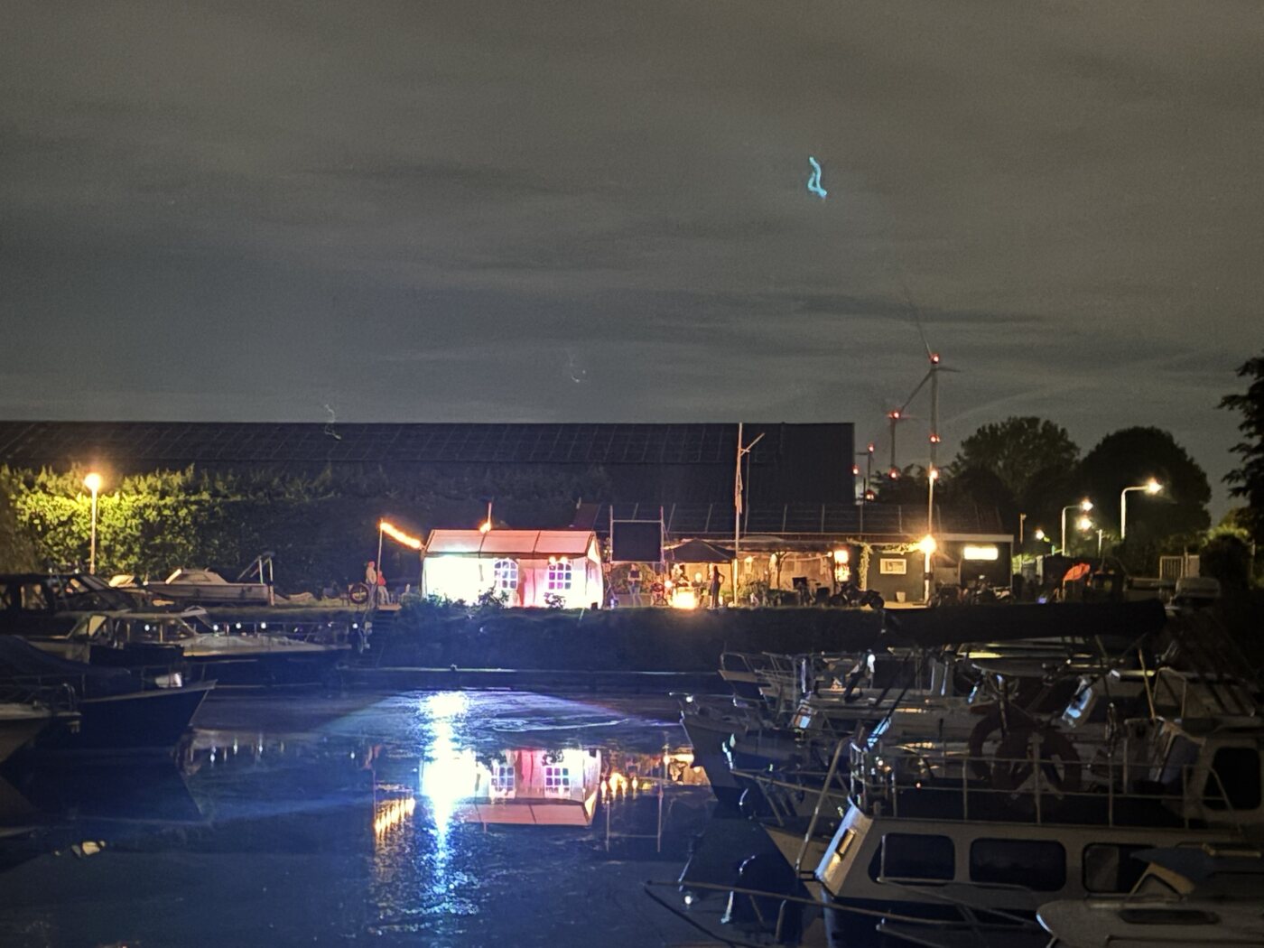 Verlicht clubgebouw en feestterrein gezien vanaf het water, tussen de aangemeerde boten